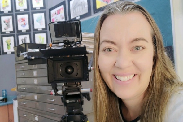 A women poses in front of a camera one tripod in a classroom. 