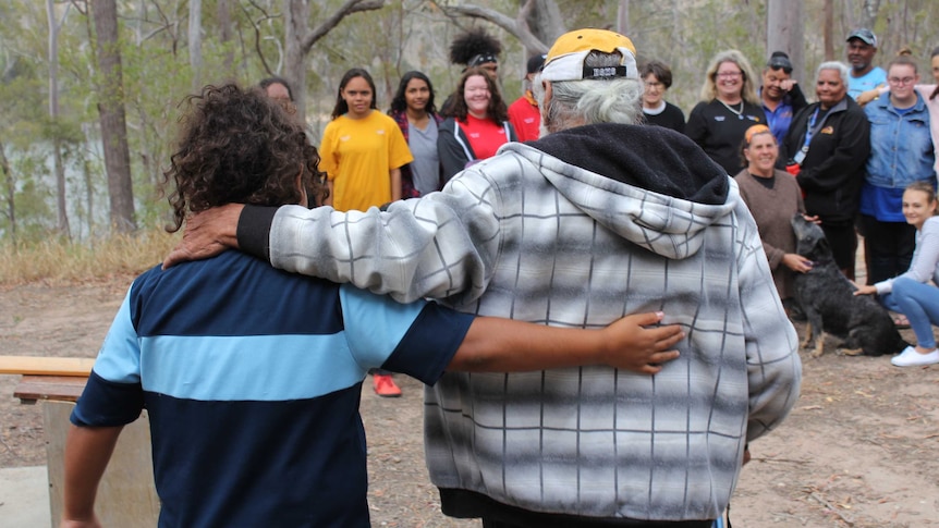 A man with grey hair and a young person with curly black hair walk arm in arm towards a group of people.