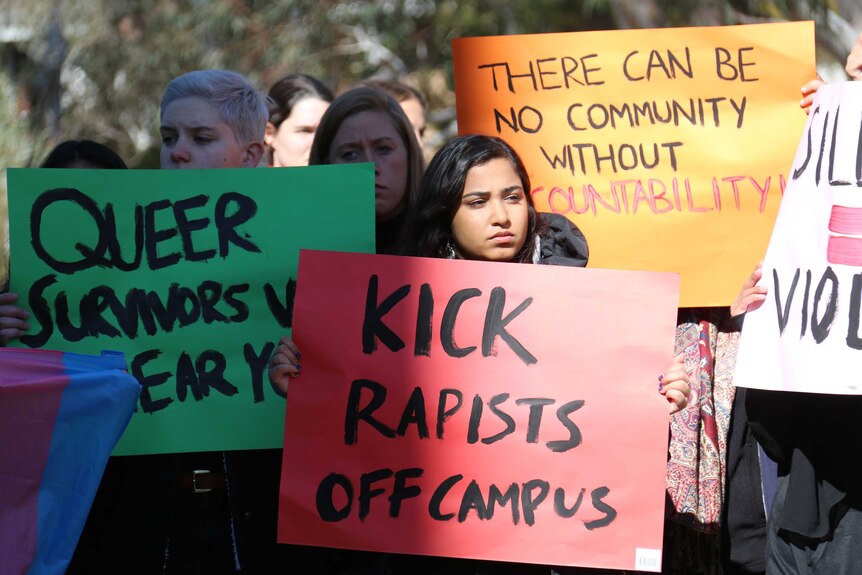 Protesters hold up signs reading 'Kick rapists off campus' and 'There can be no community without accountability'