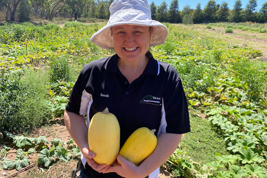 A woman stands in a pumpkin patch, smiling and holding two spaghetti squash.