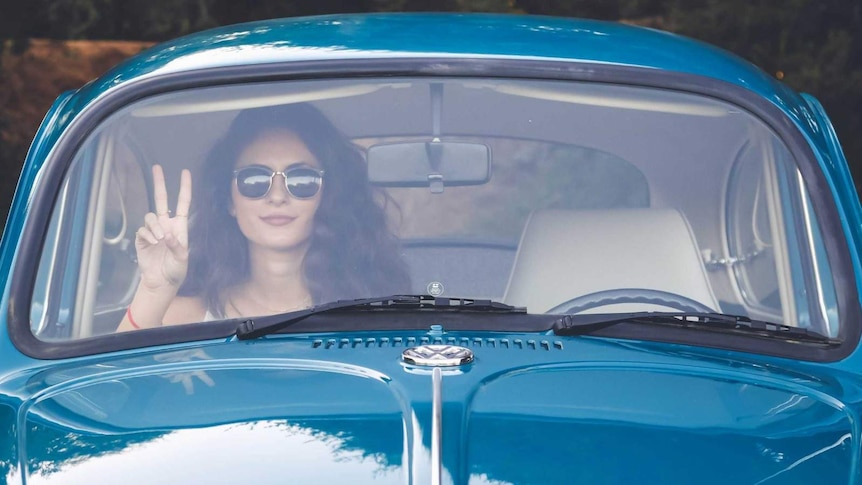 A woman sits in a blue car making the peace sign with her fingers.