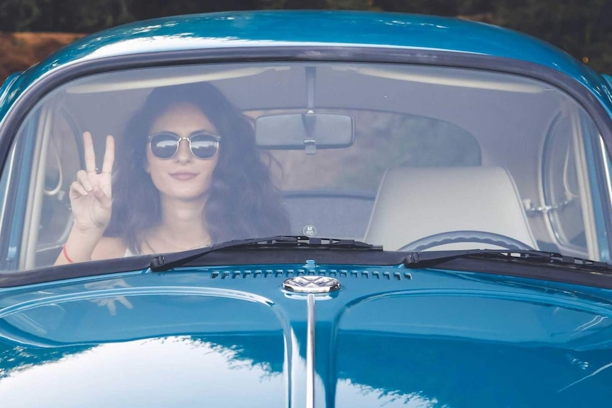 A woman sits in a blue car making the peace sign with her fingers.