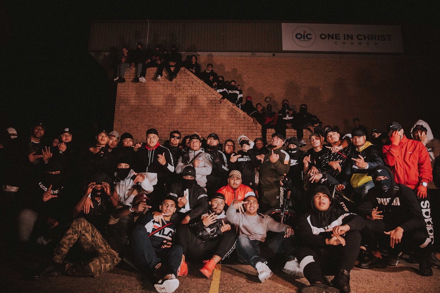 A group of young people pose together, many of pacific island appearance, they are wearing tracksuits and designer brands.