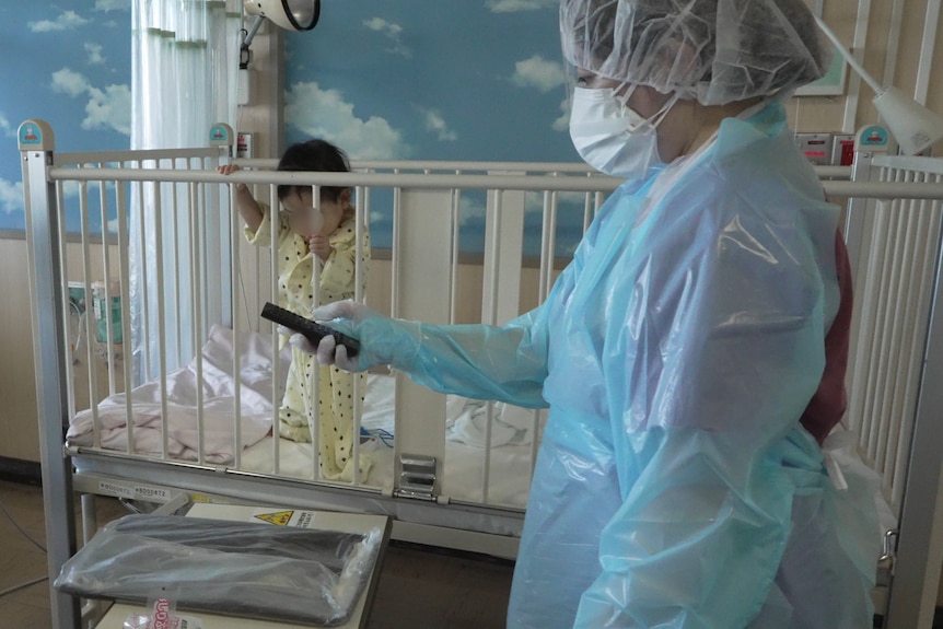 A woman in full PPE points a remote while a small baby girl stands in a cot beside her