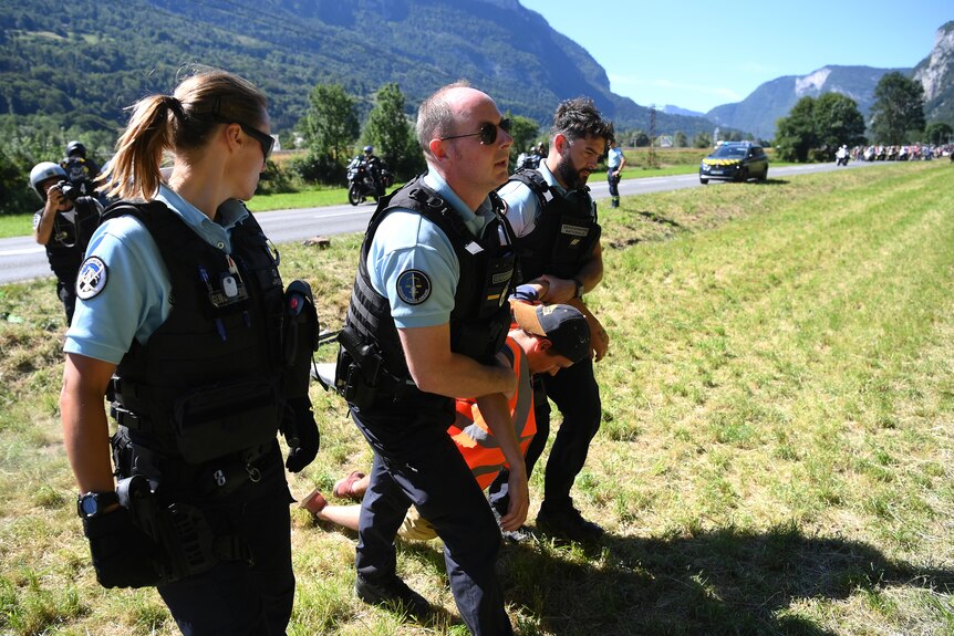 Police drag a man between them holding his shoulder as the peloton waits in the background