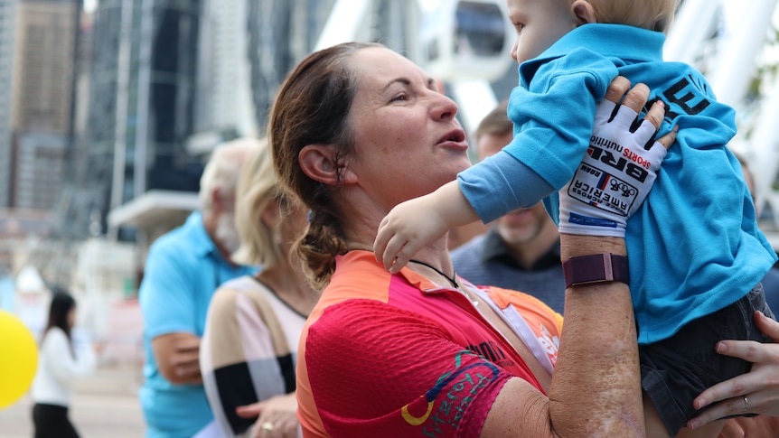 A woman in a cycling suit smiles with joy as she holds a young child above her face.