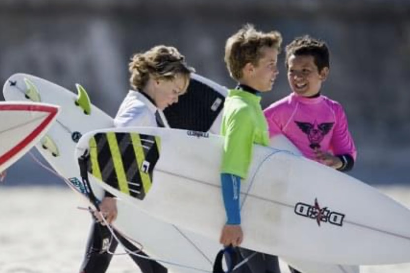 three kids hold surfboards walking on the beach.