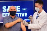 A man in a blue shirt receives an injection from a man in a white coat.