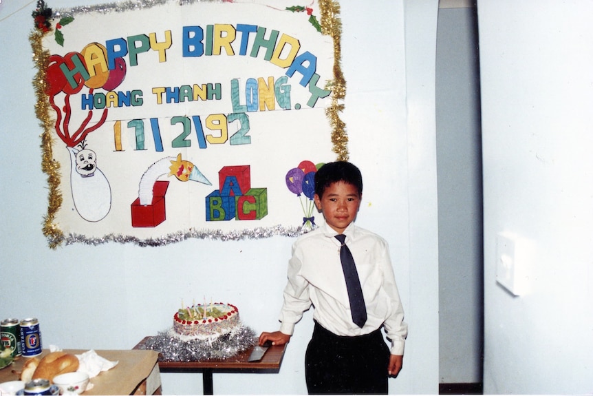 Young boy, wearing white shirt and black tie, in front of birthday poster, and next to birthday cake. 