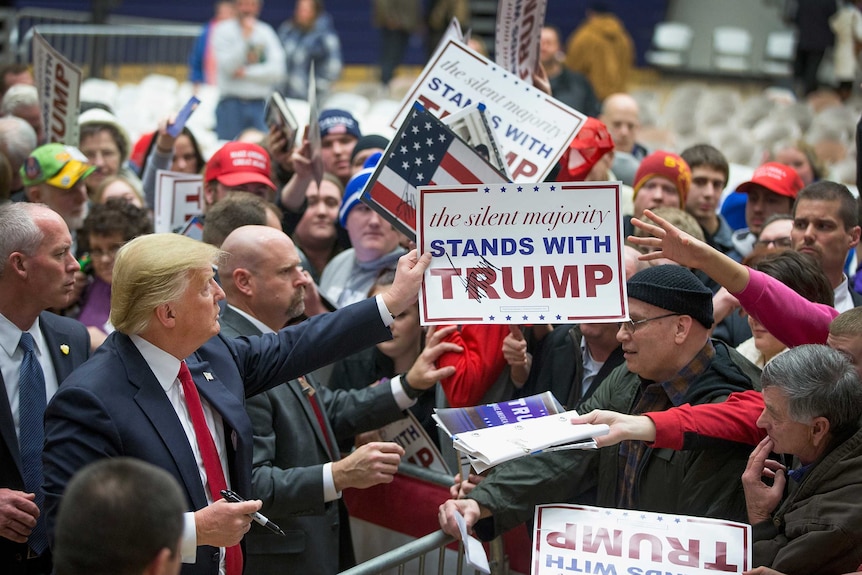Donald Trump stands with a crowd - he hands one a sign with his signature reading Trump stands with the silent majority