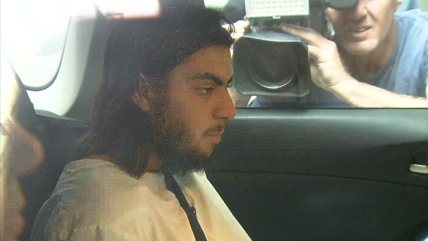 A bearded man dressed in a white gown sits in the back seat of a car as photographers film him through the passenger window.