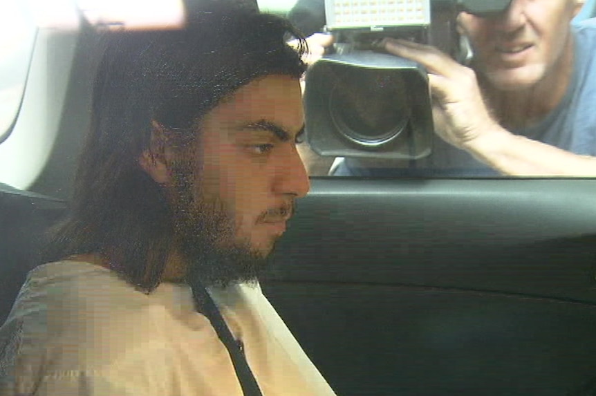 A bearded man dressed in a white gown sits in the back seat of a car as photographers film him through the passenger window.