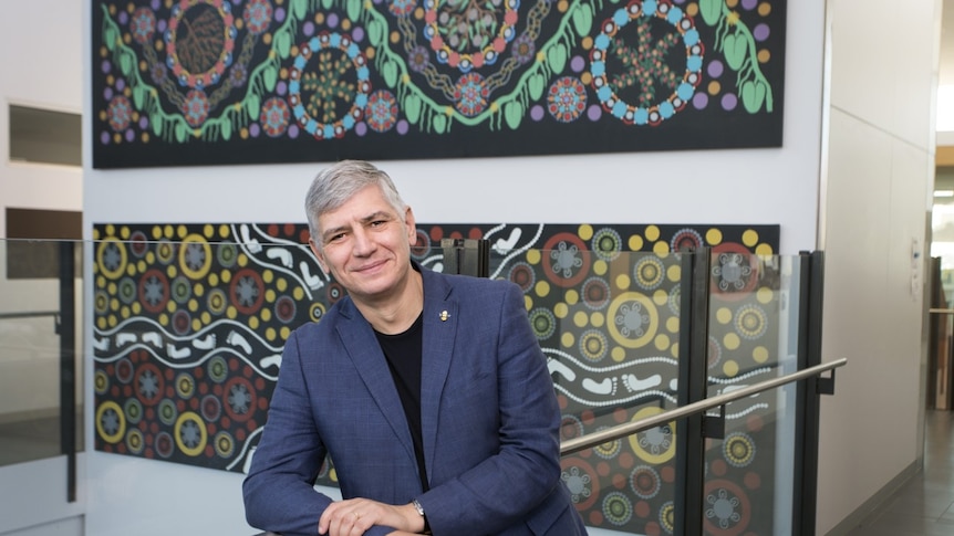 Mike Kyrios smiling at the camera with Aboriginal artwork hanging behind him