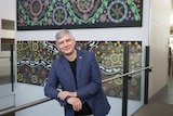 Mike Kyrios smiling at the camera with Aboriginal artwork hanging behind him