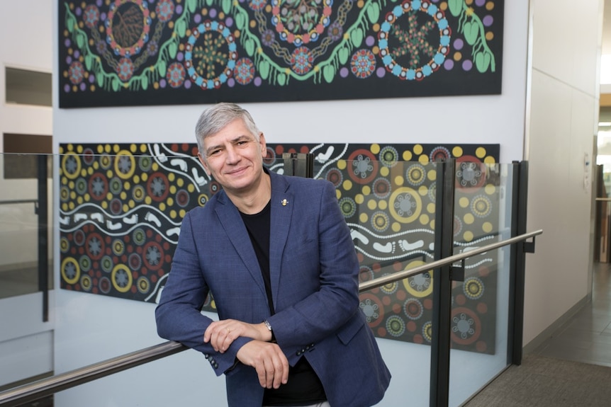 Mike Kyrios smiling at the camera with Aboriginal artwork hanging behind him