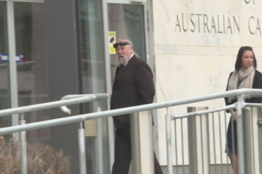 A man with a grey handlebar moustache and wearing sunglasses on his head walks into the court building.