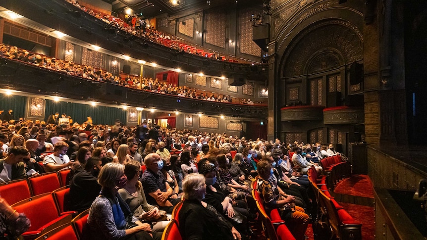side shot of mask wearing audience in large, opulent tiered theatre. red velvet seats