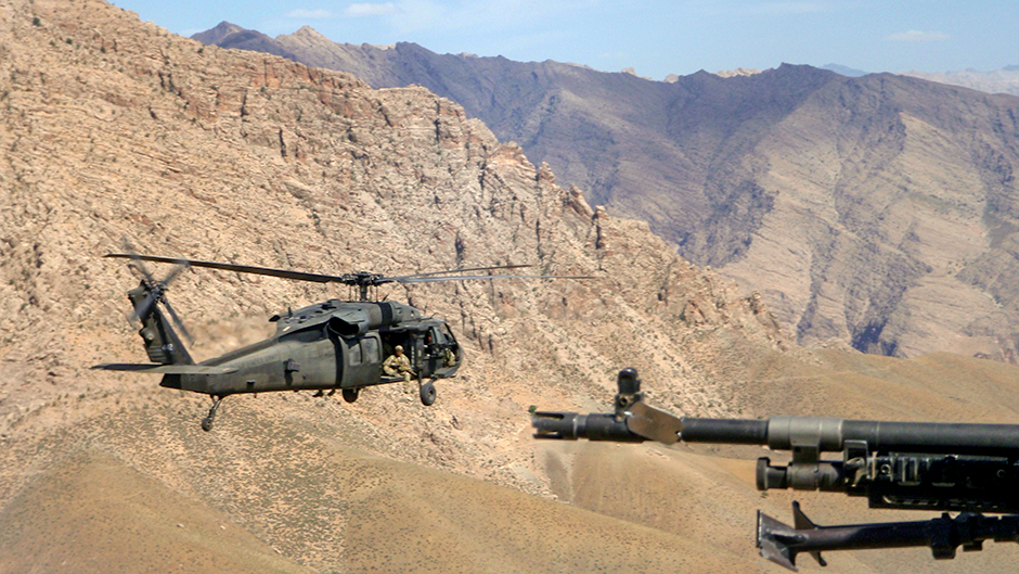 View of out of a helicopter, with gun in foreground, second helicopter and mountain range in distance