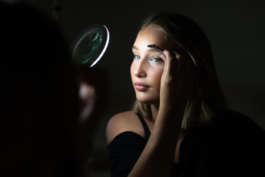 A teenager in a dark room, applying make-up in a small mirror.