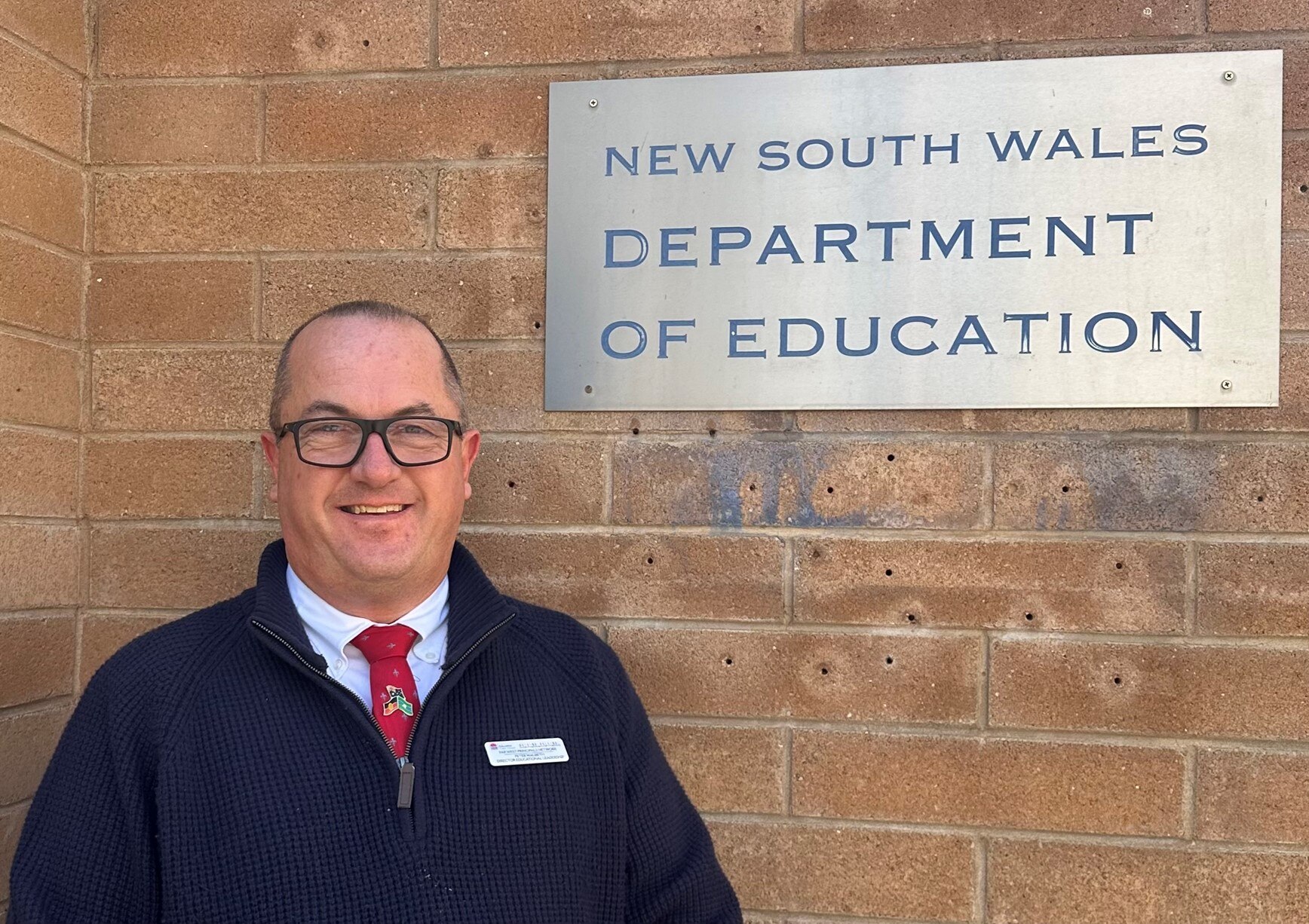 A bald man in glasses wearing a dark jumper and tie standing in front of a wall with a sign