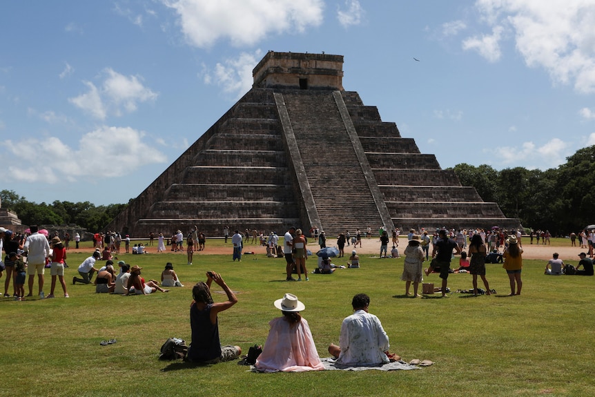 People sitting in front of a pyramid at Chichen Itza, looking up at the sky