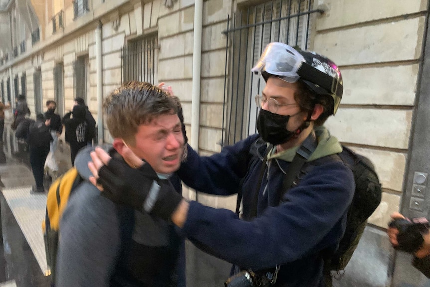 A man grimaces and is helped by another man wearing a helmet, mask and goggles.