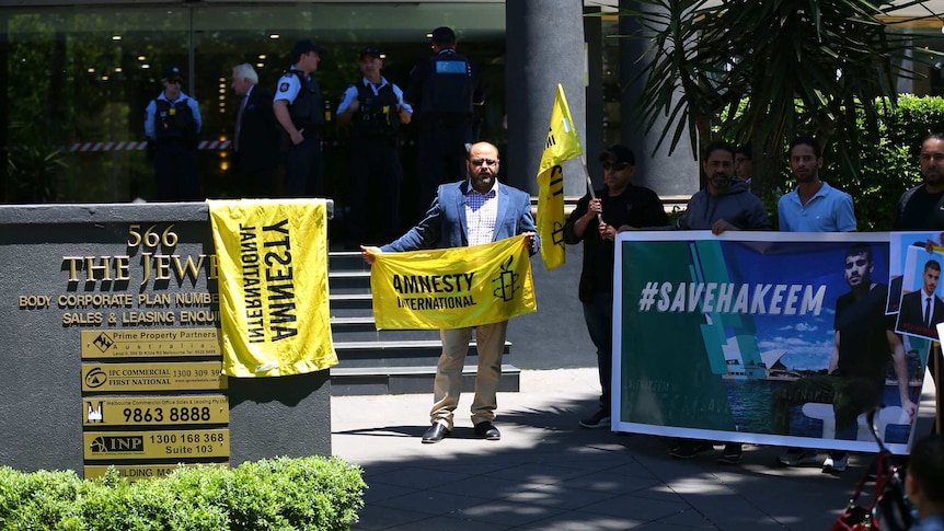 A man in a navy blazer holds an Amnesty International Flag outside the Thai consulate in Melbourne, with police in background.