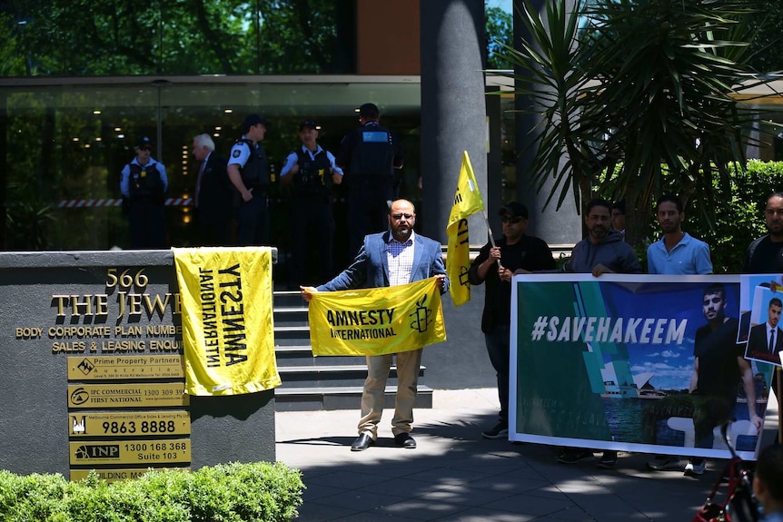 A man in a navy blazer holds an Amnesty International Flag outside the Thai consulate in Melbourne, with police in background.