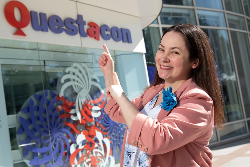 A woman points at the Questacon sign, smiling.