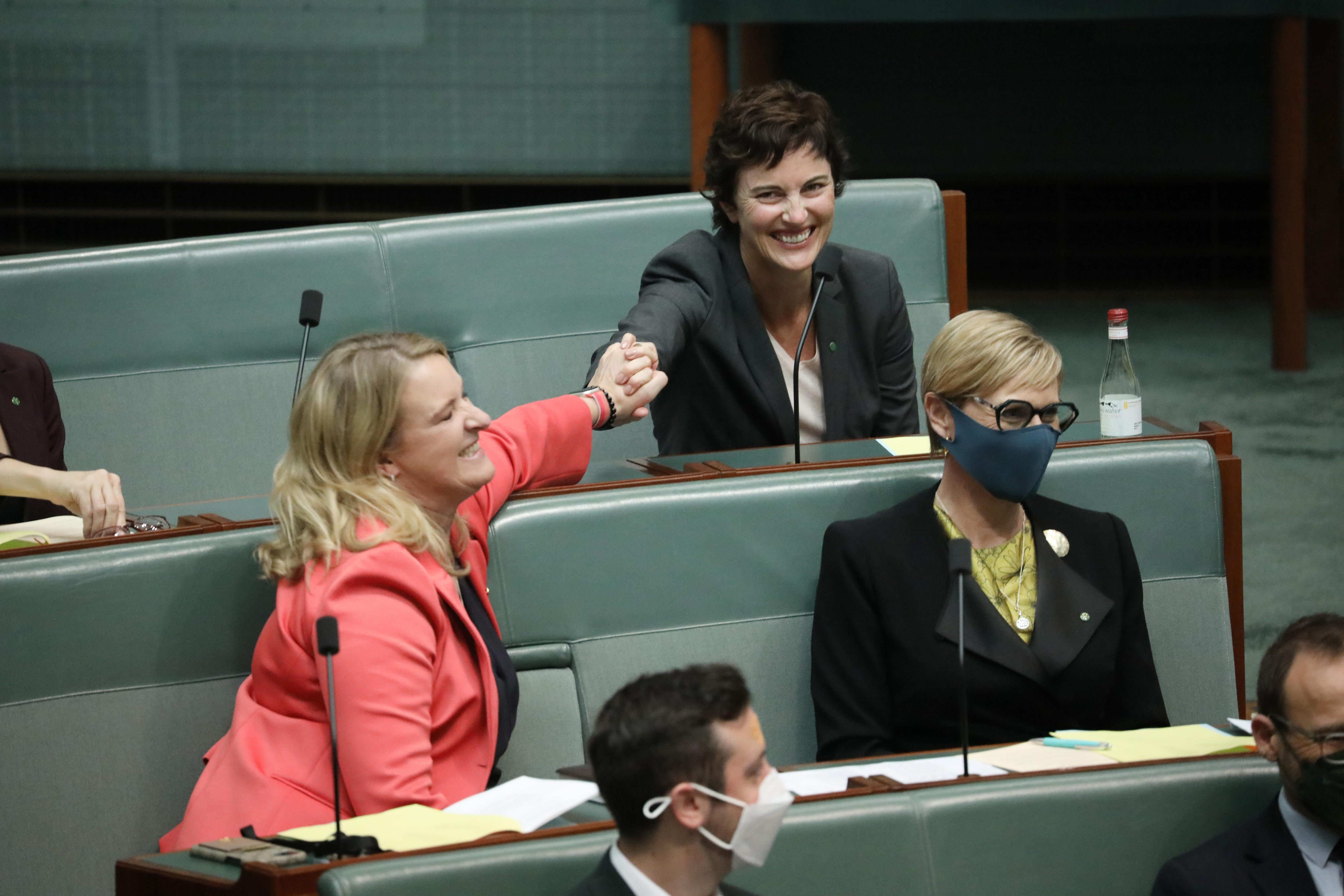 Tink and Chaney hold hands and smile while sitting in the rows of the House of Representatives.