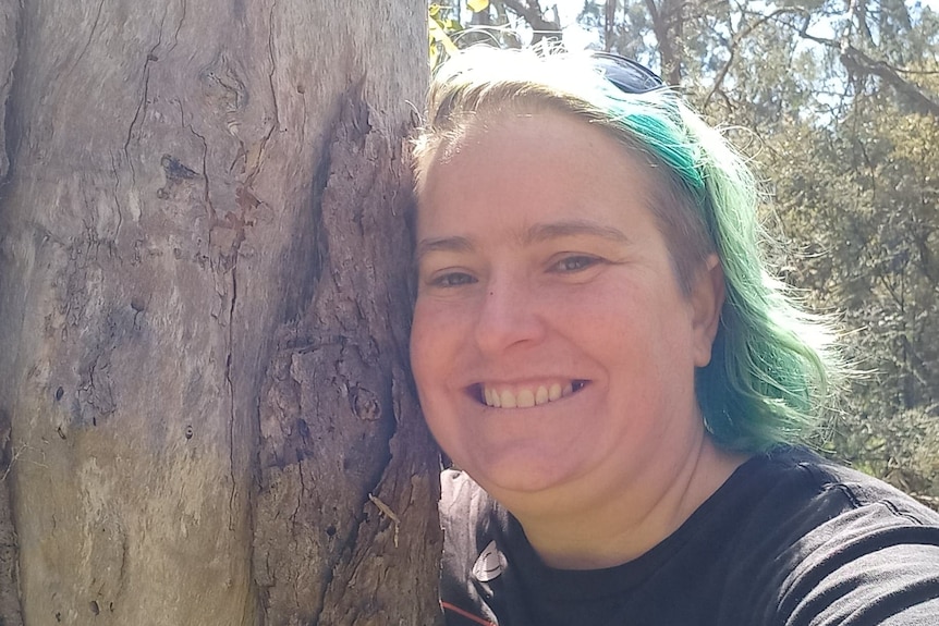A person with blue hair is seen smiling as they stand next to a tree trunk. They wear a black shirt.
