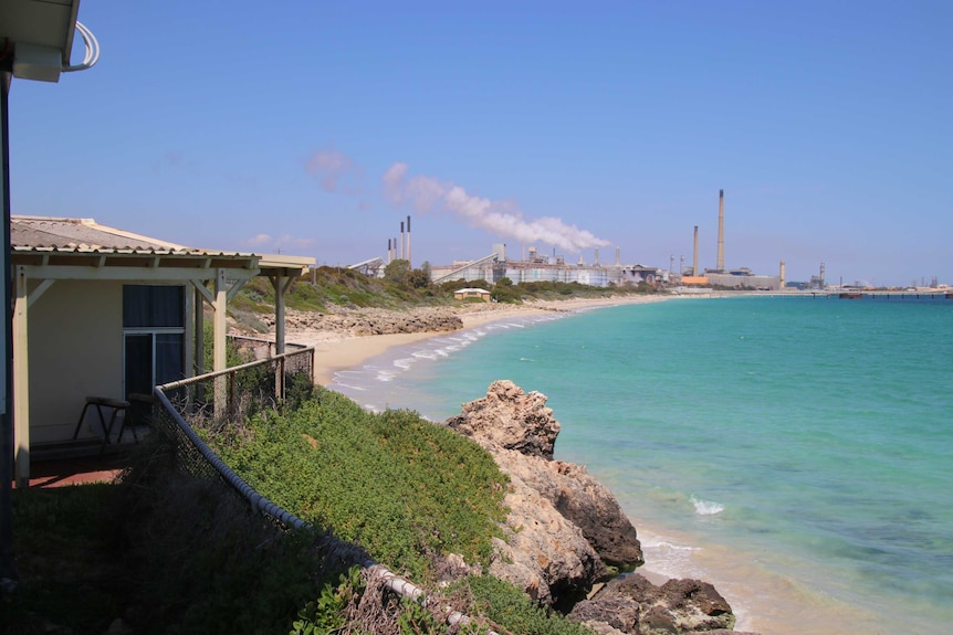 A view from the back veranda of holiday shacks facing an empty beach leading to a heavy industrial area with large smoke plumes