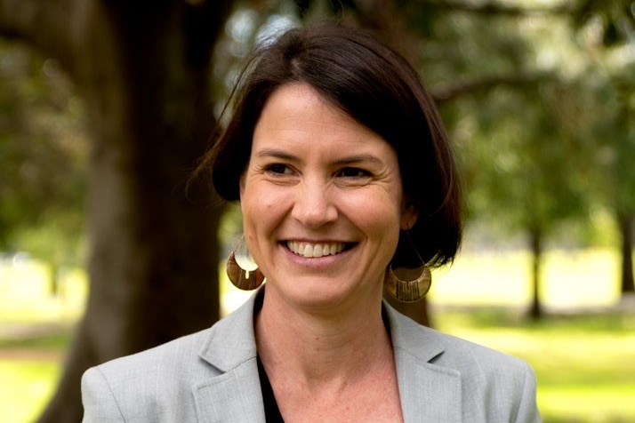 A woman with brown hair wearing a grey blazer and black top in a park