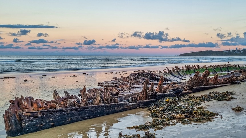 Half submerged hull of shipwreck with bits of timber poking out from sand with a sunset in the background