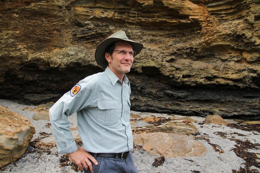 A man in a park ranger's uniform stands in front of a rock formation on a beach.