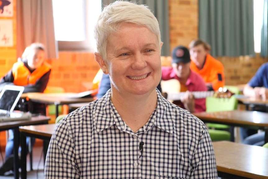 A woman with short blonde hair and a checked shirt pictured in a classroom with students in the background.