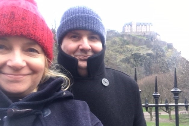 Desiree and Anthony smile in a selfie, wearing beanies in front of Edinburgh Castle.