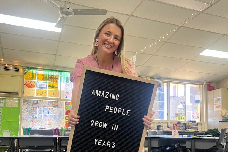 A smiling blonde woman holds sign that reads "Amazing people grow in year 3".