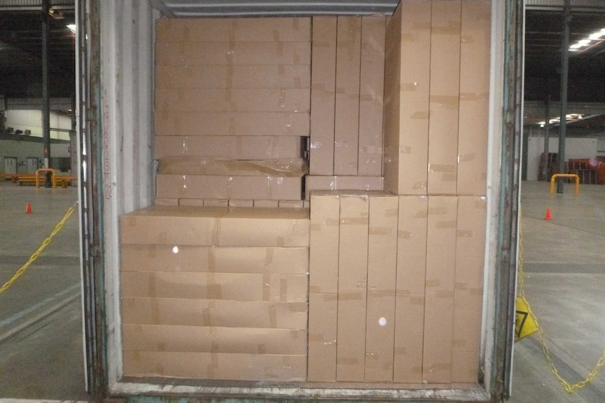 A sea container sits inside a large warehouse packed full of brown cardboard boxes.