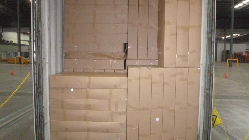 A sea container sits inside a large warehouse packed full of brown cardboard boxes.
