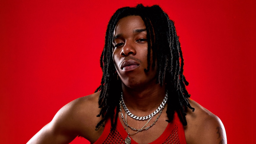 Gold Fang sits in front of a red backdrop in a red mesh singlet and silver necklaces