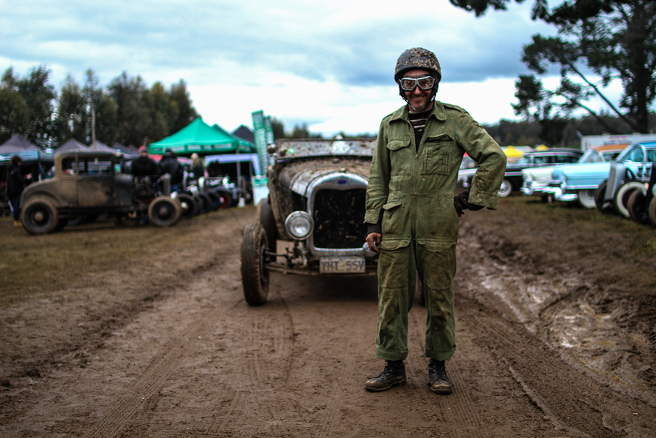 A man in green overalls, goggles and helmet in front of an old topless car, on a muddy road.