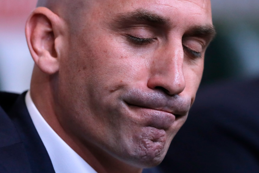 A bald man in a suit looks serious with his eyes closed in close-up.