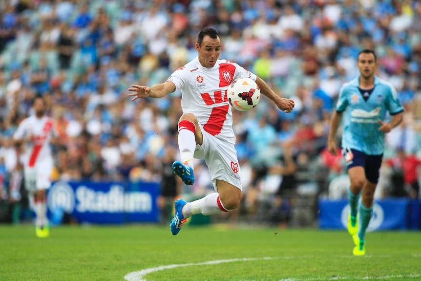 An A-League footballer leaps in the air to trap the ball during a game.