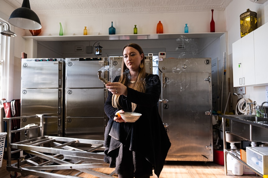  a woman wearing all blacks holds a smudge stuck to sage a mortuary room, containing coloured glass jars top stainless steel fre