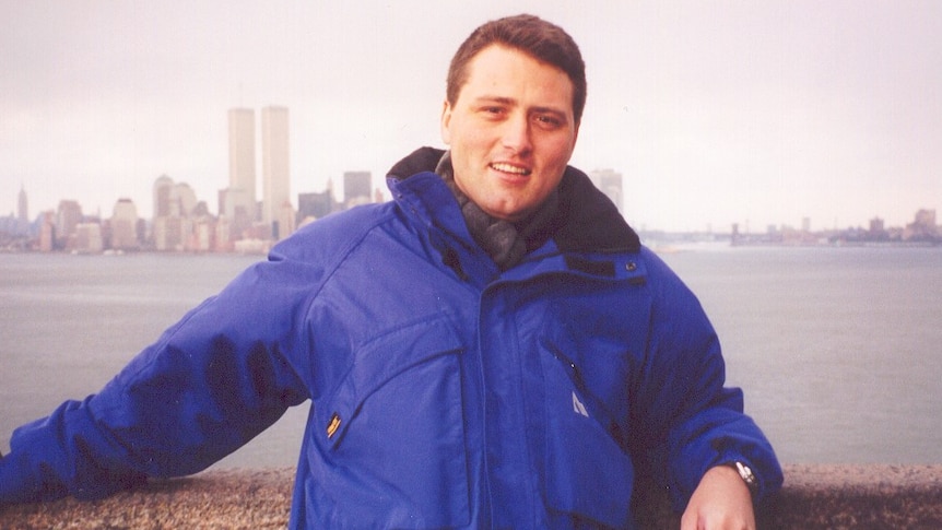 A man wearing a blue coat standing in front of a river, with the original World Trade Center in the background