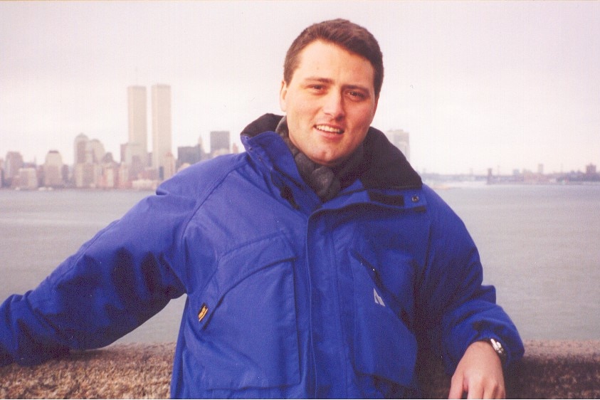A man wearing a blue coat standing in front of the East River and the World Trade Center