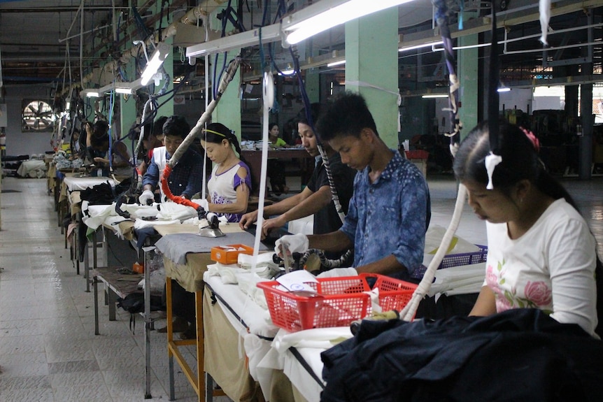 A man, and two women workers iron garments on tables in a long row in a factory.