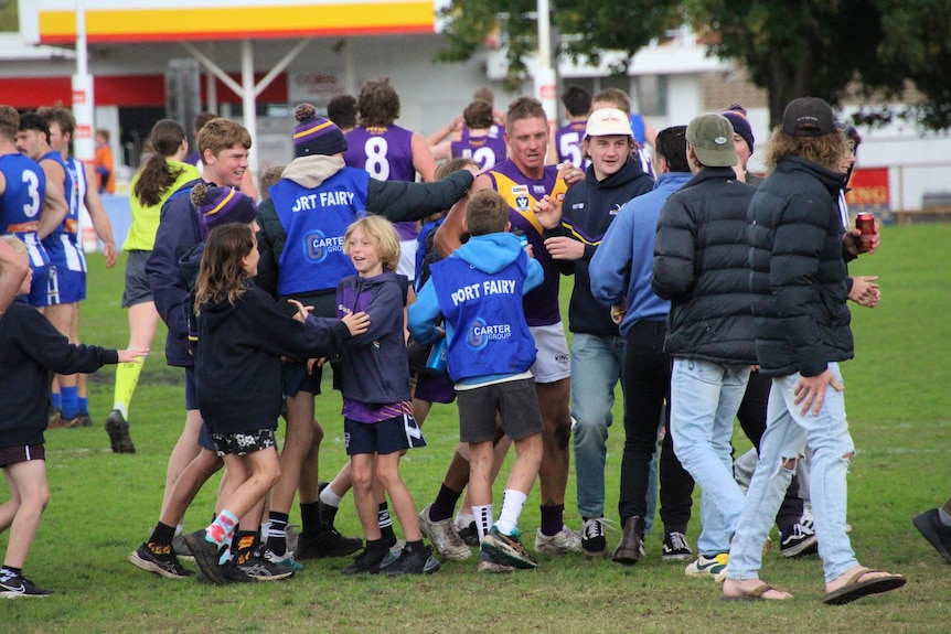 Young boys and teenagers crowd around the football player on the oval. 