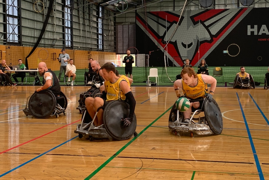 A red-haired wheelchair rugby player pushes his chair hard to make ground while carrying the ball in his lap.